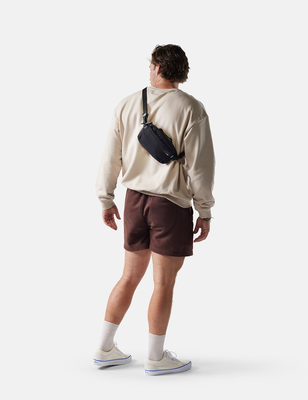 Man wearing a beige sweater, brown shorts, and carrying a black bag on a white background